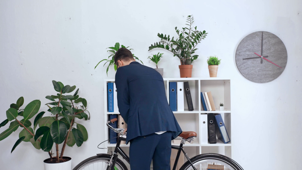 handsome businessman carrying bike in office and placing it near rack with folders - Footage, Video