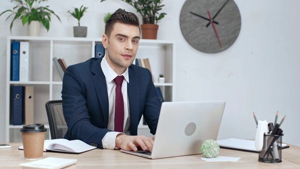 good-looking businessman checking time at wristwatch, smiling and looking at camera while using laptop at workplace - Materiał filmowy, wideo