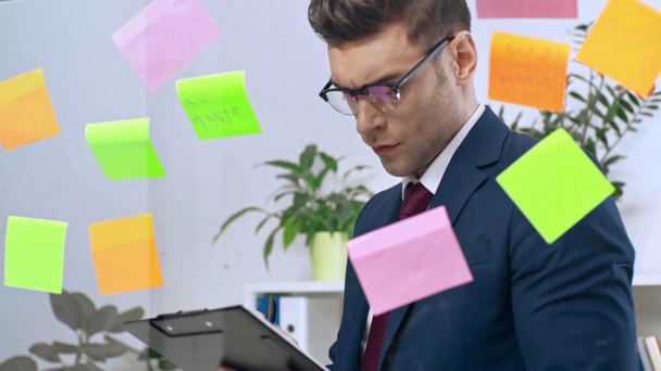 attentive businessman looking at colorful sticky notes and writing in clipboard - Кадры, видео