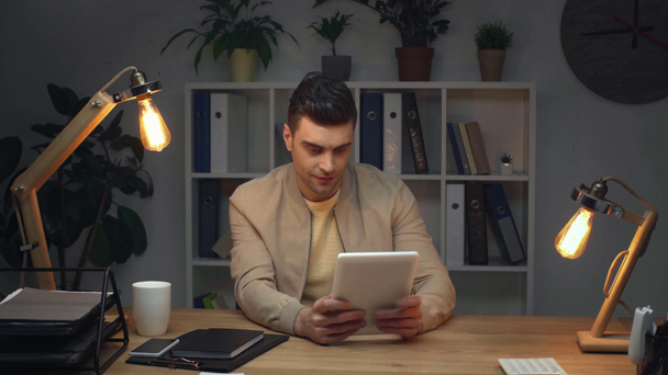uomo d'affari sorridente utilizzando tablet digitale mentre seduto sul posto di lavoro in ufficio buio di notte
 - Filmati, video