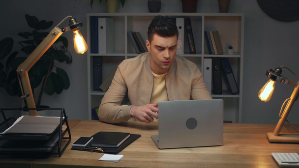 bonito empresário beber café e usando laptop enquanto sentado no local de trabalho no escritório à noite
 - Filmagem, Vídeo