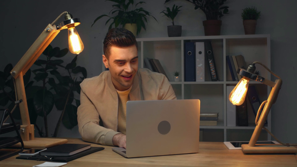 excited businessman showing yes gesture while using laptop at workplace in dark office at night - Imágenes, Vídeo