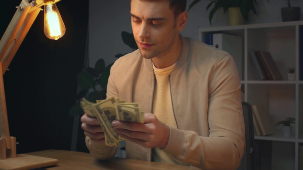 uśmiechający się biznesmen liczenia banknotów dolara siedząc przy biurku pracy w biurze w nocy - Materiał filmowy, wideo
