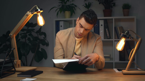 handsome, serious businessman writing in clipboard while sitting at workplace in office at nighttime - Imágenes, Vídeo