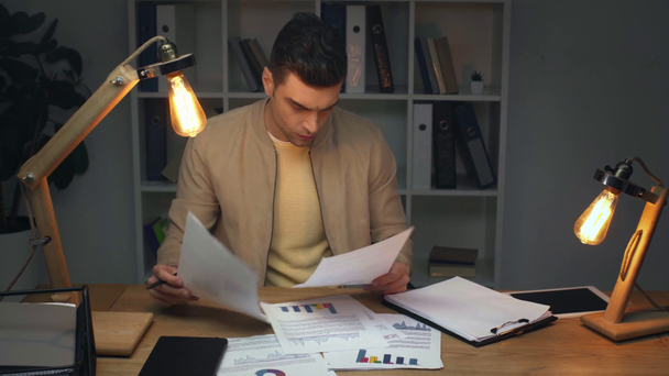 attentive businessman studying document with graphs and charts and writing in clipboard while sitting at workplace in office at night - Filmati, video