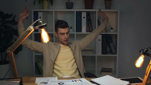 tired businessman studying document with charts and graphs, writing in clipboard, stretching and yawning while sitting at workplace in office at night - Video, Çekim