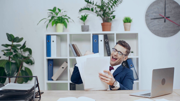 Zeitlupe eines glücklichen Geschäftsmannes, der am Schreibtisch im Büro Dokumente wirft und Ja-Geste zeigt - Filmmaterial, Video