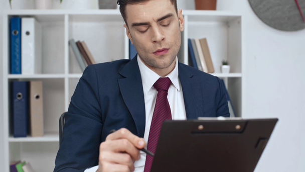selective focus of thoughtful businessman writing in clipboard in office - Metraje, vídeo