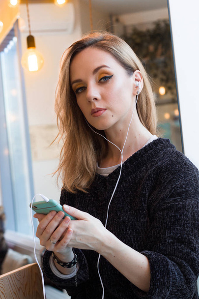 beautiful smiling young woman in gray sweater using mobile phone with earphones in cafe - Photo, image
