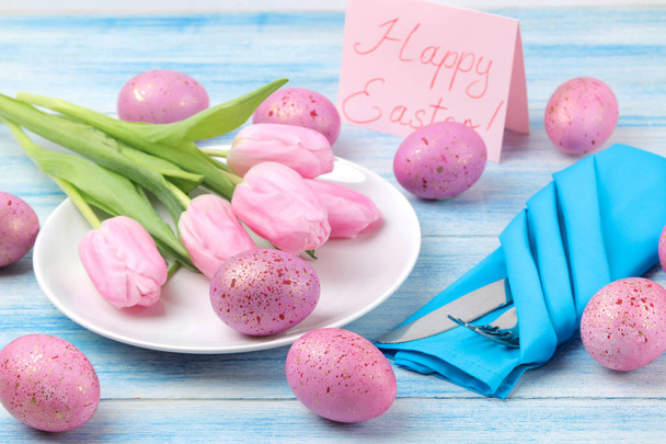 Пасха. Накрытие пасхального стола. Розовые пасхальные яйца и цветы тюльпаны на голубом деревянном столе. Счастливой Пасхи. праздники
. - Фото, изображение