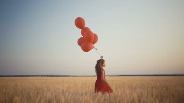 šťastná mladá dívka s balóny, které jsou spuštěny v poli pšenice při západu slunce. 4k video. - Záběry, video