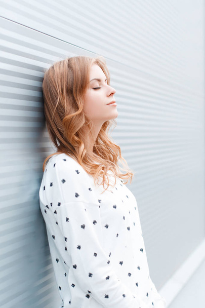 Krásná mladá žena se světlými vlasy ve stylově bílém pulovníku, který odpočívá u moderní kovové zdi ve městě. Rozkošný Evropský dívčí model relaxuje v teplém jarním dnu. - Fotografie, Obrázek