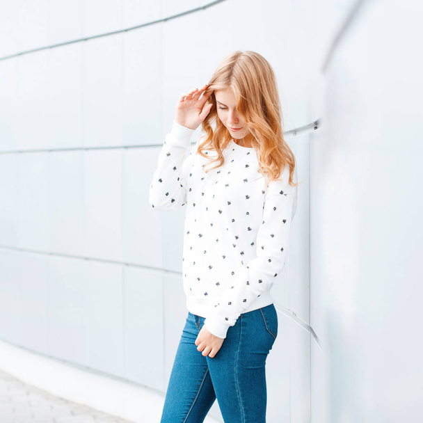 Veselou atraktivní mladou ženu s módním účesu v bílém svetru v módních džínách u moderní zdi ve městě. Krásná šťastná dívka si užívá víkend. - Fotografie, Obrázek