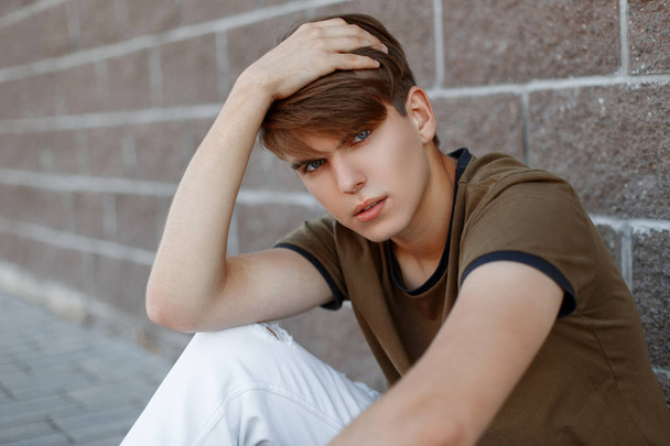 Europejski młody człowiek w modnej zielonej T-shirt w stylowe białe spodnie z modną fryzurę relaksuje siedzi w pobliżu rocznika muru ceglanego. Ładny nowoczesny model mody facet odpoczywa. - Zdjęcie, obraz