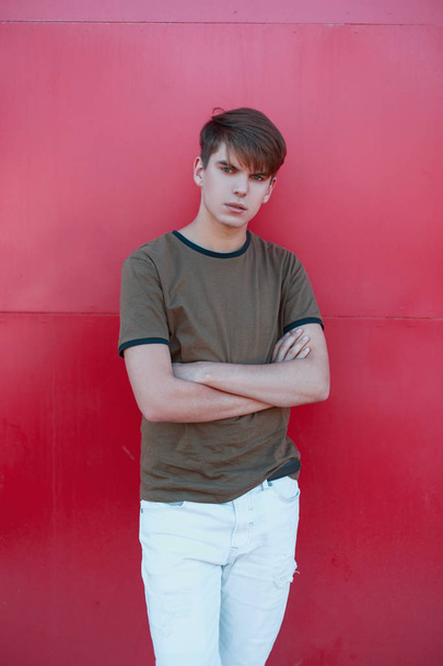 Fashionable modern handsome young man with a fashionable hairstyle in a green summer stylish T-shirt in white pants posing near a bright pink metal wall. Cool modern guy is a teenage. - 写真・画像