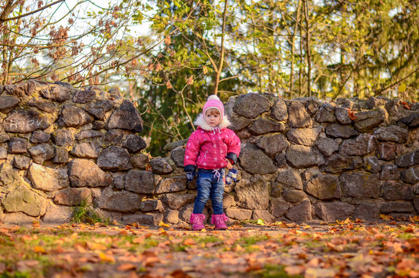  Μικρό κορίτσι σε ρούχα του χειμώνα βρίσκεται σε ιστορικό πέτρινο τοίχο - Φωτογραφία, εικόνα