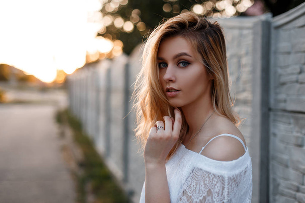 Retrato de una hermosa joven rubia europea con ojos azules con maquillaje natural en una elegante blusa de encaje blanco cerca de una valla vintage al atardecer. Linda chica en un paseo en el verano
. - Foto, imagen