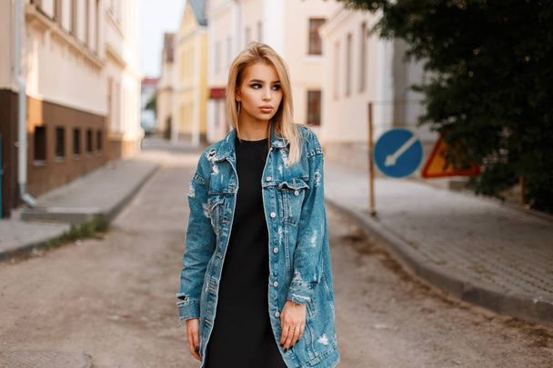 Stylish urban pretty young woman in a black stylish dress in a fashionable long denim jacket posing in a city on the street near vintage buildings. Cute girl outdoors. Spring style women's clothing. - Photo, Image