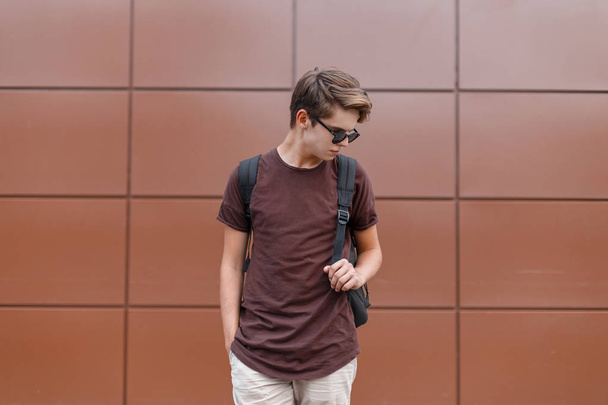 Jonge knappe hipster man met een modieuze kapsel in korte broek in zwart stijlvolle zonnebrillen in een Vintage T-shirt met een stijlvolle rugzak staat in de buurt van de Vintage muur. Cool Guy geniet van het weekend - Foto, afbeelding