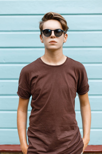 Americký stylový mladý muž s módním hnědým tričkem v černých slunečních brýlích s módním účesu v ulici nedaleko starobyle dřevěné budovy. Moderní moderák venku v letním dni. - Fotografie, Obrázek