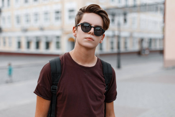 Vakava komea nuori hipster mies muodikas t-paita tyylikäs kampauksen reppu seisoo kadulla lämpimänä kesäpäivänä. Houkutteleva kaveri nauttii kävellä kaupungissa
. - Valokuva, kuva