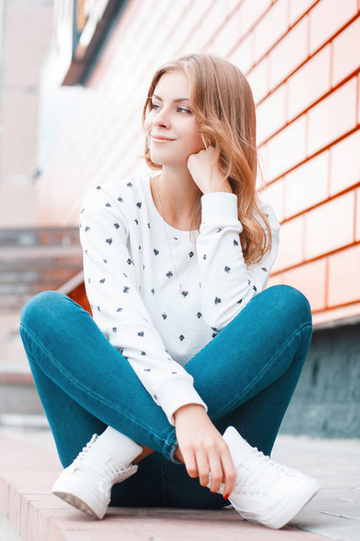 Cheerful pretty european young woman in a white sweater in fashionable jeans in sneakers sits and dreams near the modern brick building on the street. Cute blonde girl enjoys a holiday. - 写真・画像