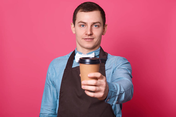 Young handsome male barista suggests you cup of coffee made by him, dressed elegant blue shirt, white bow tie and brown apron, isolated over pink background. Copy space for your logo. Drink concept. - Foto, Bild
