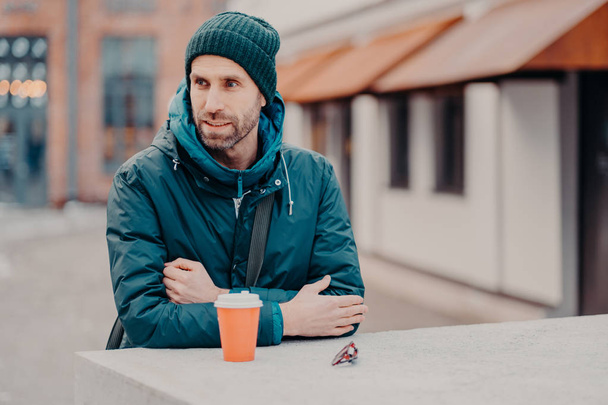Pensive unshaven young Caucasian man with bristle, wears hat and jacket with hoody, drinks takeaway coffee, stands outdoor, has stroll. People, lifestyle, spare time and street style concept. - Photo, image