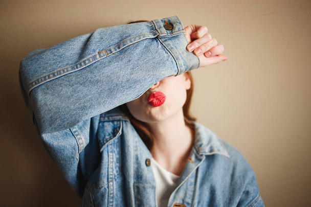 Young anonymous woman in denim jacket covering her face with her hand and showing kiss gesture - Фото, изображение