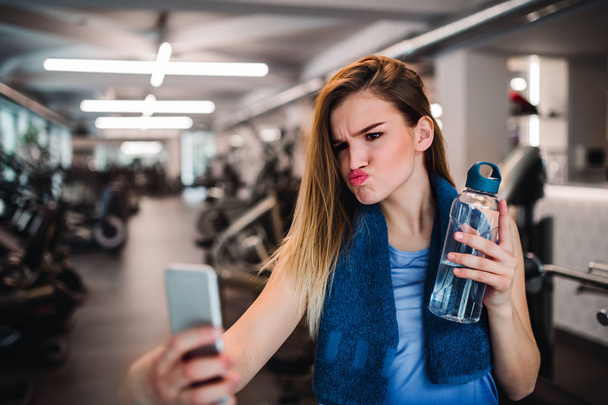 A portrait of young girl or woman with smartphone in a gym, taking selfie. - Foto, Imagen