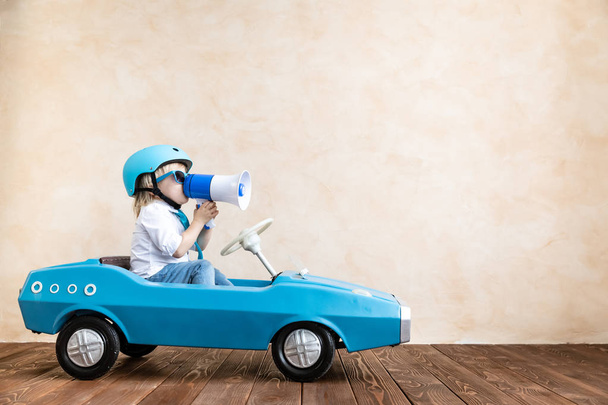 Happy child speaking to megaphone at home. Funny kid driving toy car indoor. Success and win concept - Photo, Image