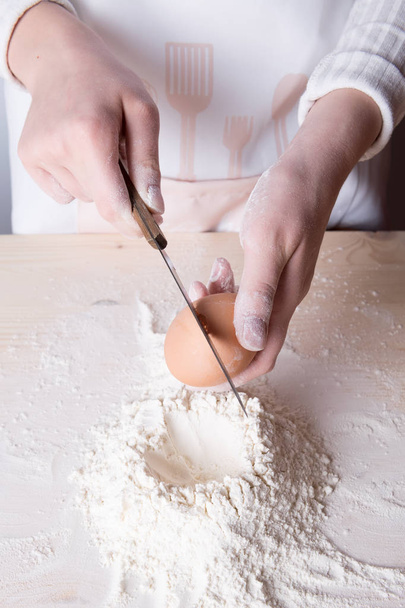 Mujer joven rompe el huevo por encima de la harina blanca para hacer masa para pasta italiana
 - Foto, Imagen