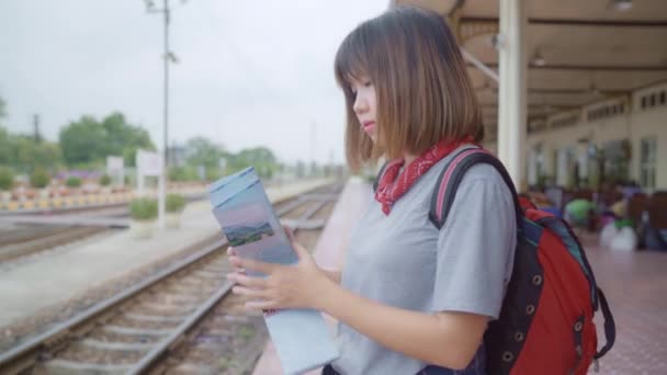 Utazó ázsiai nő irányát és látszó-on Térkép-míg a kiadások nyaralás utazás és a vasútállomáson várakozó vonat, hátizsákos fiatal női turista élvez a utazás. Életmód nők utazási koncepció - Felvétel, videó