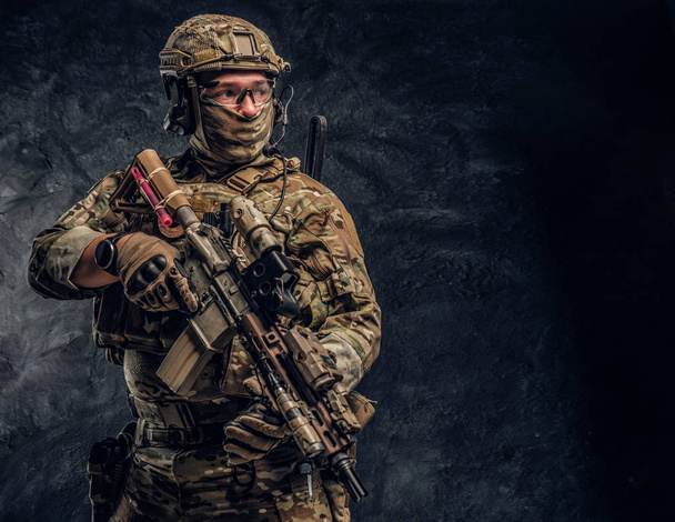 Volledig uitgeruste soldaat in camouflage uniforme houdt een aanvalsgeweer. Studio foto tegen een donkere muur - Foto, afbeelding