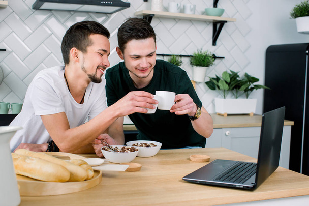 Two young handsome men having breakfast and drinking coffee while using laptop in the kitchen at the morning. Smiling men watching film using laptop and having breakfast - Valokuva, kuva