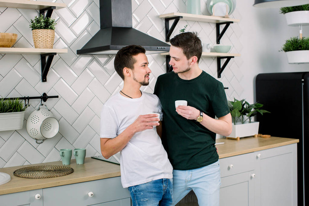 Happy gay couple with coffee at home kitchen, closeup. Handsome homosexual men drinking coffee and looking each other at the kitchen - Photo, Image