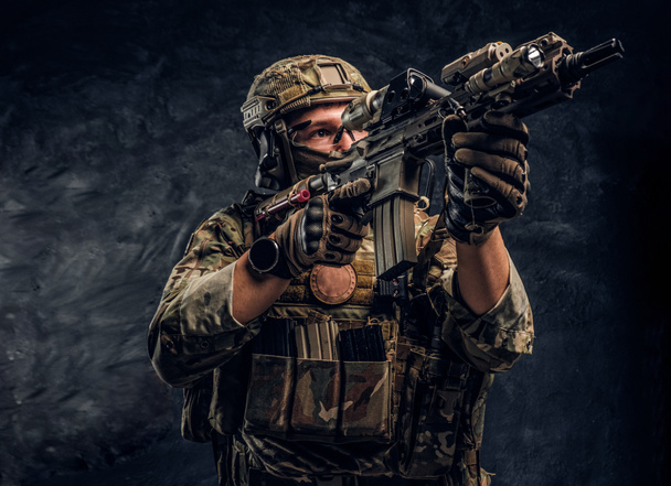 Το ελίτ μονάδα ειδικών δυνάμεων στρατιώτη με τη στολή παραλλαγής κρατώντας ένα τουφέκι επιθέσεων με ένα θέαμα με λέιζερ και στοχεύει στο στόχο. Στούντιο φωτογραφία ενάντια σε έναν τοίχο σκούρο   - Φωτογραφία, εικόνα