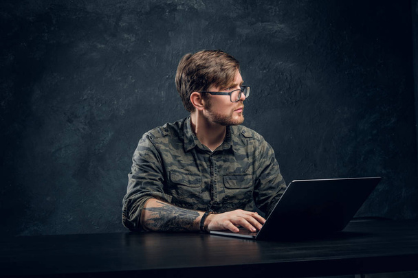 Programmeur dragen van een trendy militaire shirt zitten aan de tafel met laptop in Bureau tegen een donkere muur - Foto, afbeelding