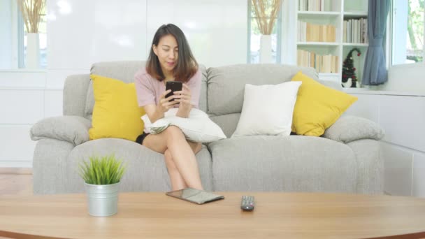 Jovem mulher asiática usando smartphone verificando redes sociais sentindo-se feliz sorrindo enquanto deitado no sofá quando relaxar na sala de estar em casa. Estilo de vida latina e hispânica etnia mulheres em casa conceito. - Filmagem, Vídeo