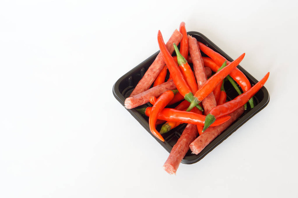 Κόκκινες καυτές πιπεριές τσίλι και λεπτά βαυαρικά λουκάνικα βρίσκονται σε μαύρη βάση σε λευκό φόντο. Καρύκευμα και υγιεινά τρόφιμα.  - Φωτογραφία, εικόνα