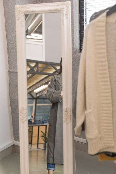 Дзеркало в магазині одягу, в ньому відображення дівчини в сірому пальто
 - Фото, зображення
