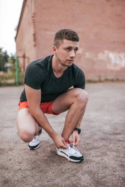 Athlete man crouched, tying shoelaces on sneakers. In the summer on the sports ground in the city. Trainer guy in the watch tracker bracelet after jogging. Workout outdoor lifestyle. - 写真・画像