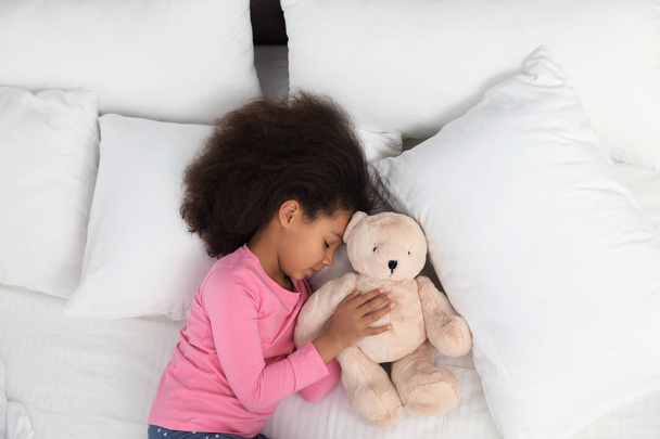 Cute little African-American girl with teddy bear sleeping in bed, top view - Foto, imagen