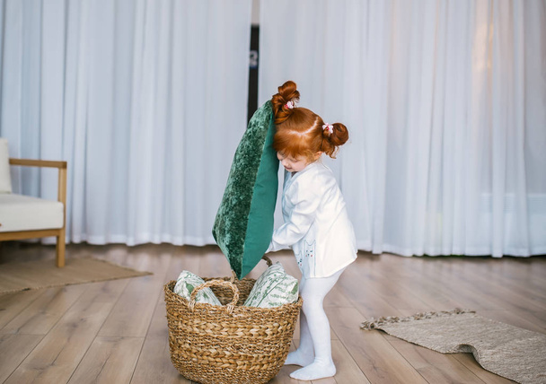 Menina ruiva feliz em pijama jogar com cesta e travesseiros no chão em casa
. - Foto, Imagem