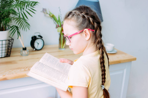 Lány szemüveges és Pigtailek olvasás könyv az asztalnál a szobájában - Fotó, kép