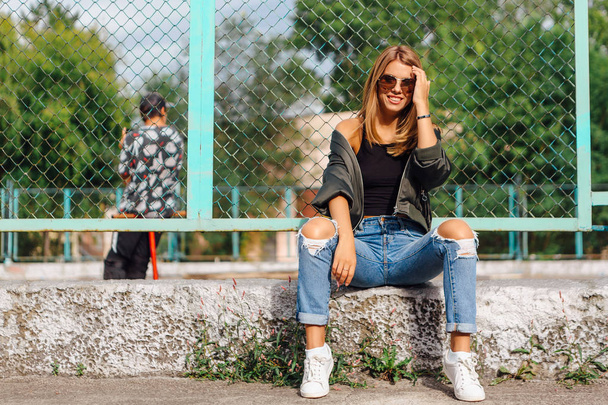 Güneş gözlüğü ve sıva şehrin yanında oturan bombardıman ceket giyen trendy genç kadın moda portre - Fotoğraf, Görsel
