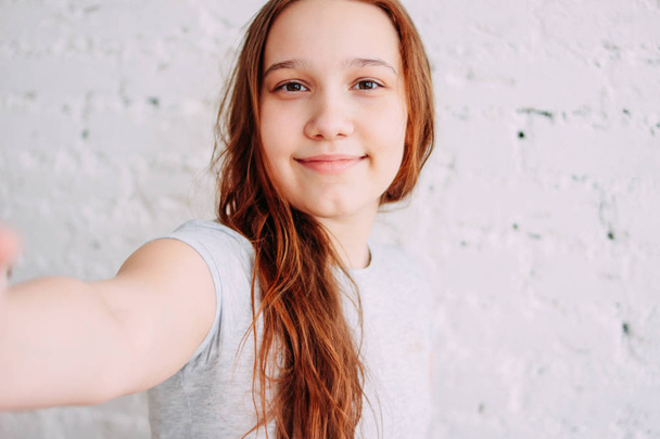 Mooie charmante Redhead glimlachende tiener meisje met selfie op frontale camera geïsoleerd op witte bakstenen muur - Foto, afbeelding