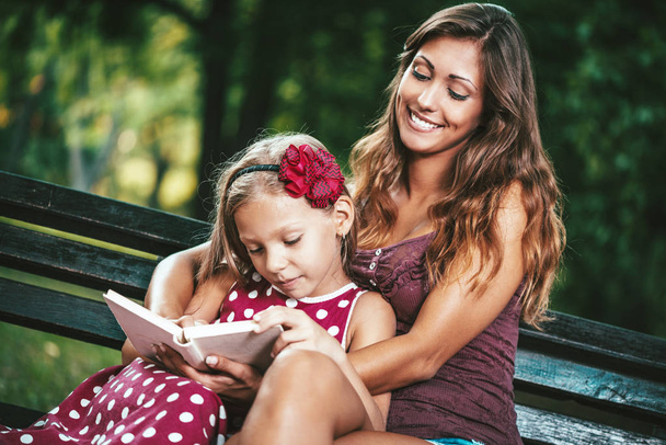little girl with mother reading book at bench in park - Φωτογραφία, εικόνα