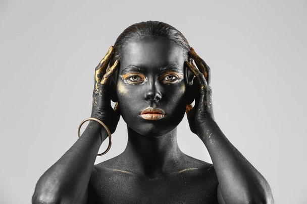 Mooie vrouw met zwarte en gouden verf op haar lichaam tegen de lichte achtergrond - Foto, afbeelding