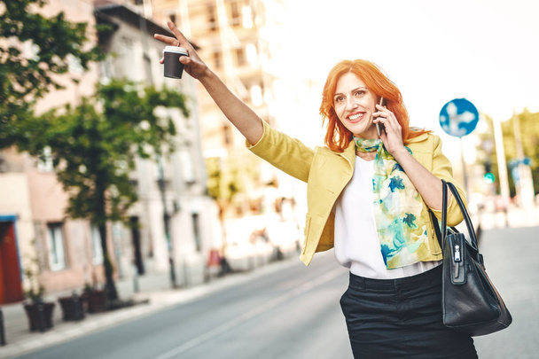 Smiling business woman standing on street with smartphone and calling for taxi - Foto, Bild